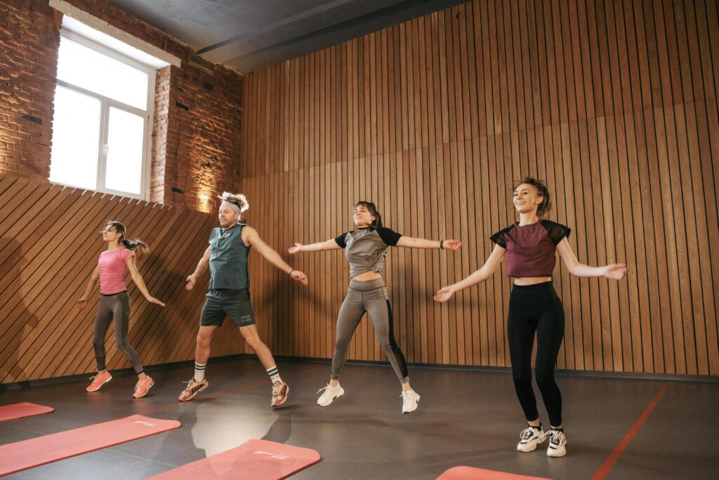 Four people performing jumping jacks in an open space gym.