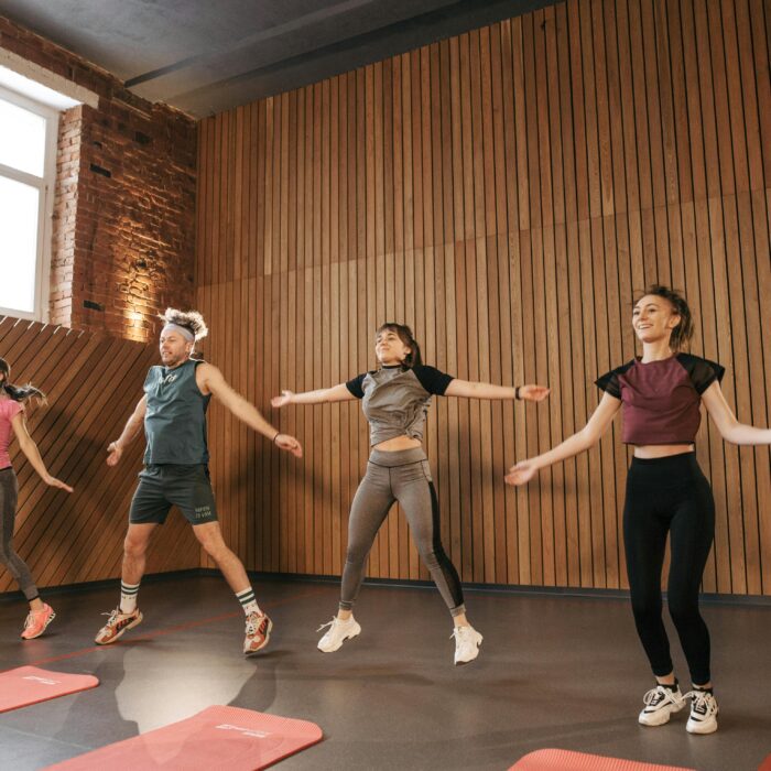 Four people performing jumping jacks in an open space gym.