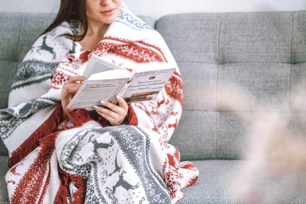 A woman wrapped in a blanket, sitting on a cozy settee, reading a book