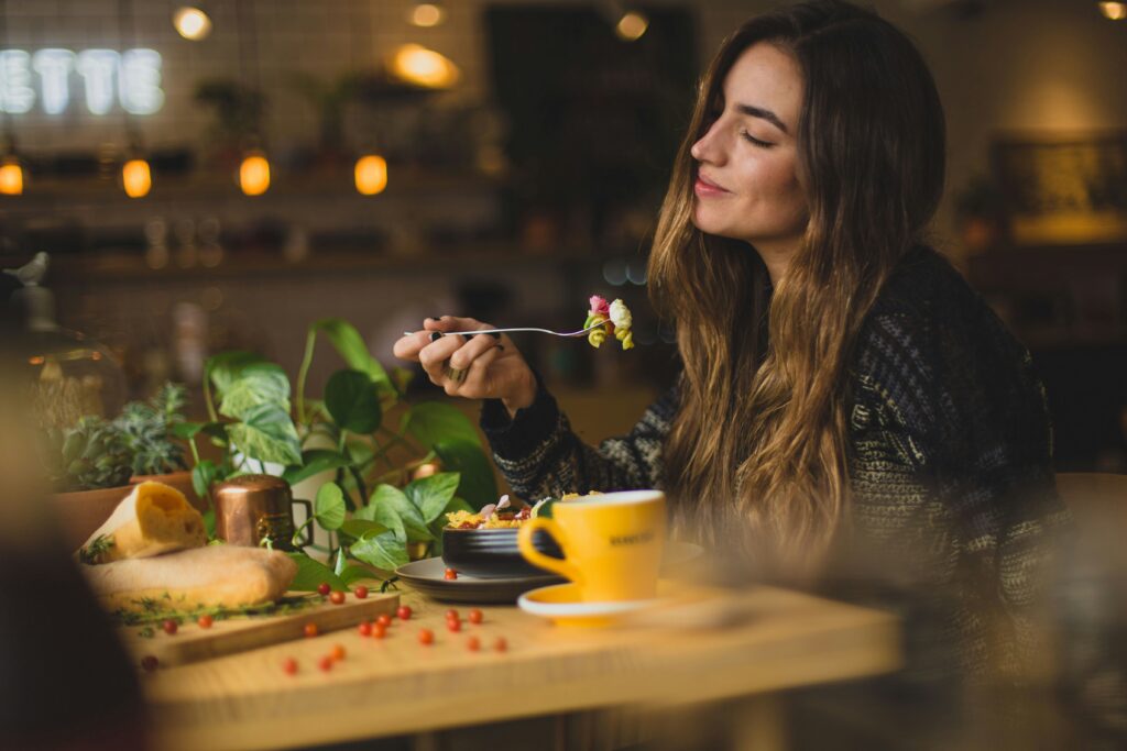 A happy woman sitting at a table, about to eat with food on her fork.