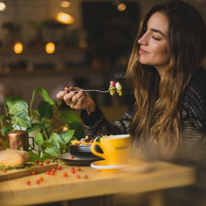 A happy woman sitting at a table, about to eat with food on her fork.