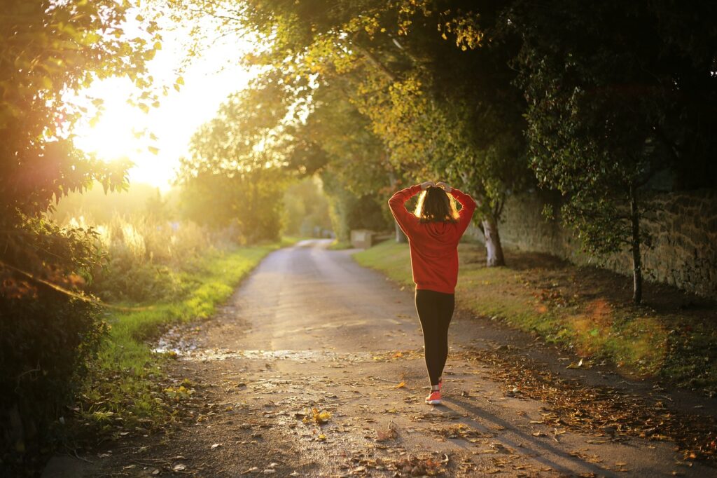 A woman walking outside in autumn with the bright sun in the background and trees lining both sides of the path