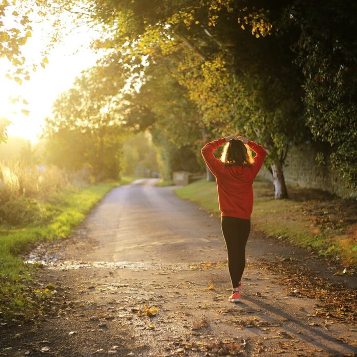 A woman walking outside in autumn with the bright sun in the background and trees lining both sides of the path