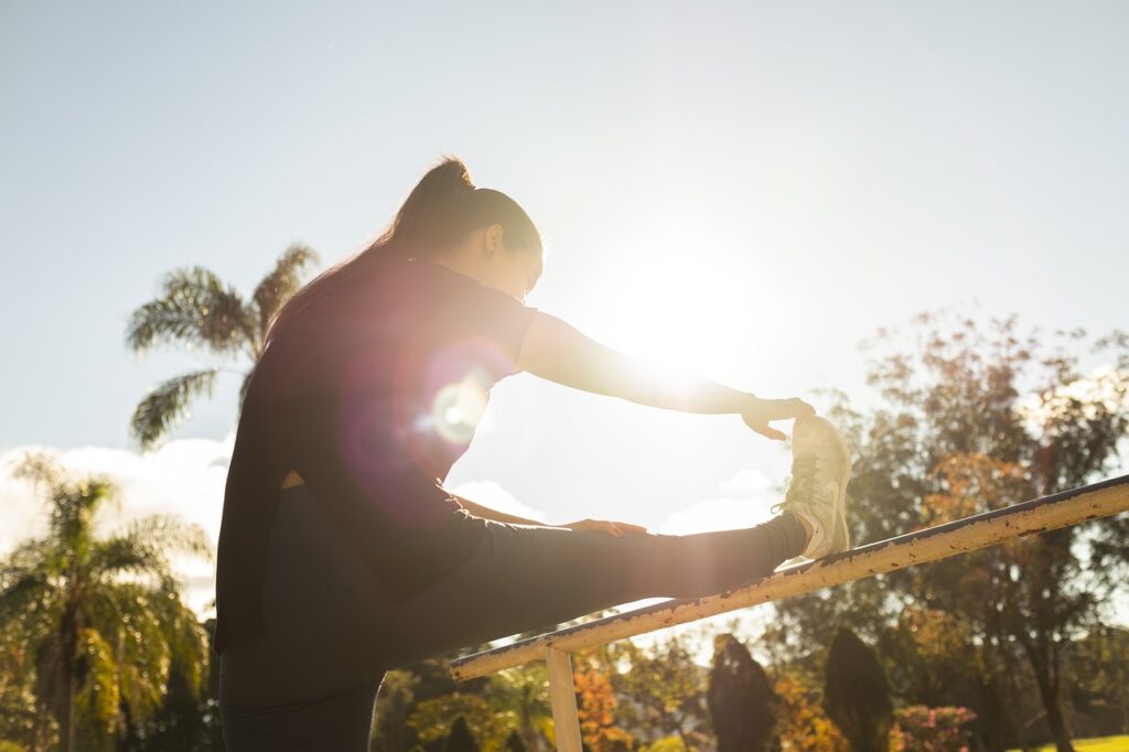 A woman outdoors, lifting one leg onto a railing and stretching, with an open sky in the background.